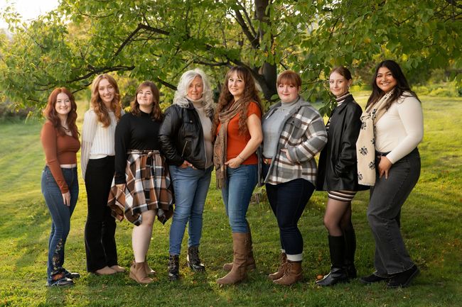 Group of eight women standing in a park, smiling at the camera. Fall attire in warm sunlight.