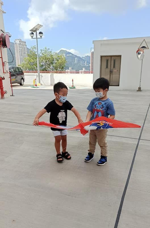Two young boys wearing face masks are flying a kite in a parking lot.