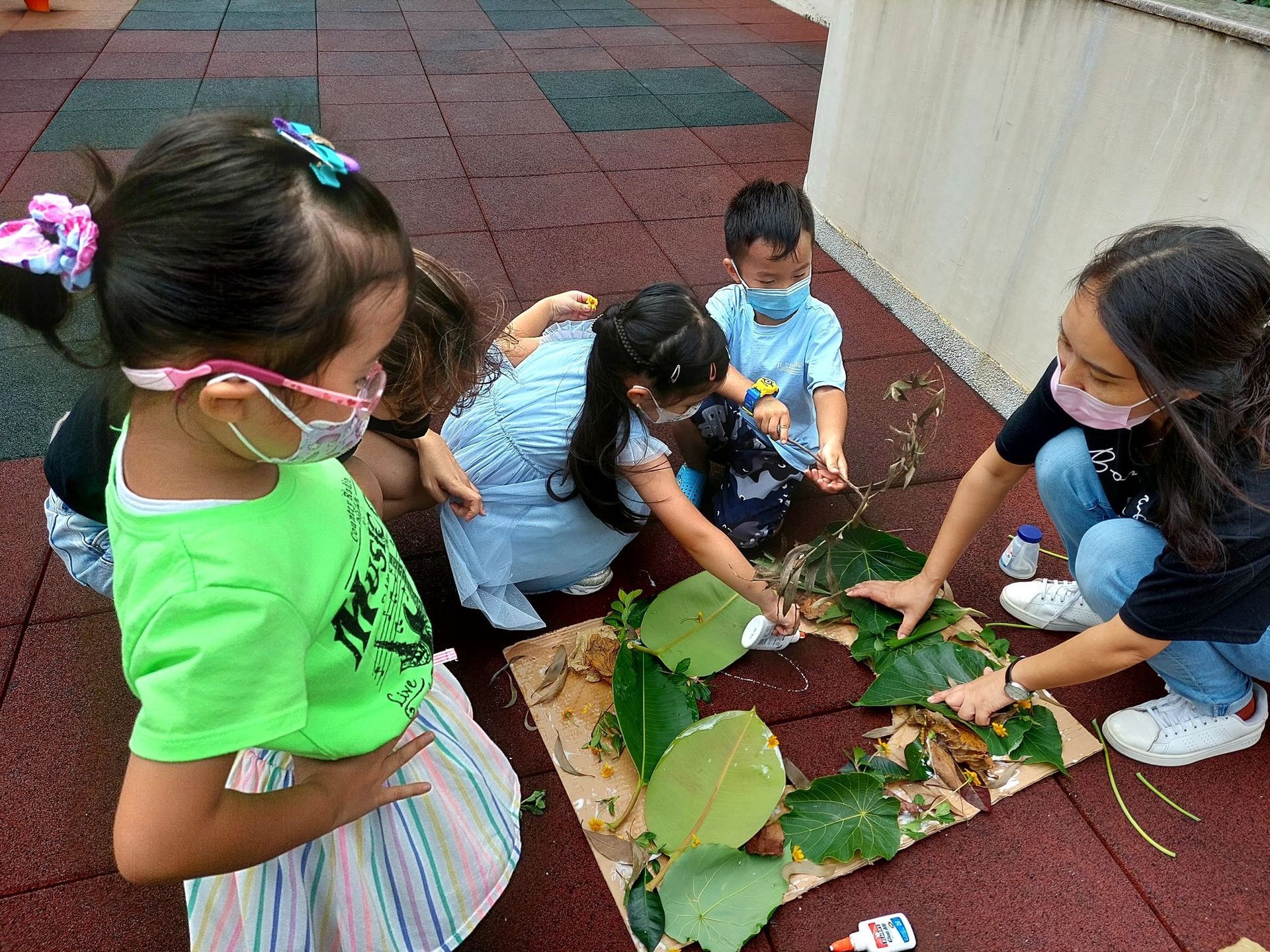 A group of children wearing face masks are playing with leaves on the ground.