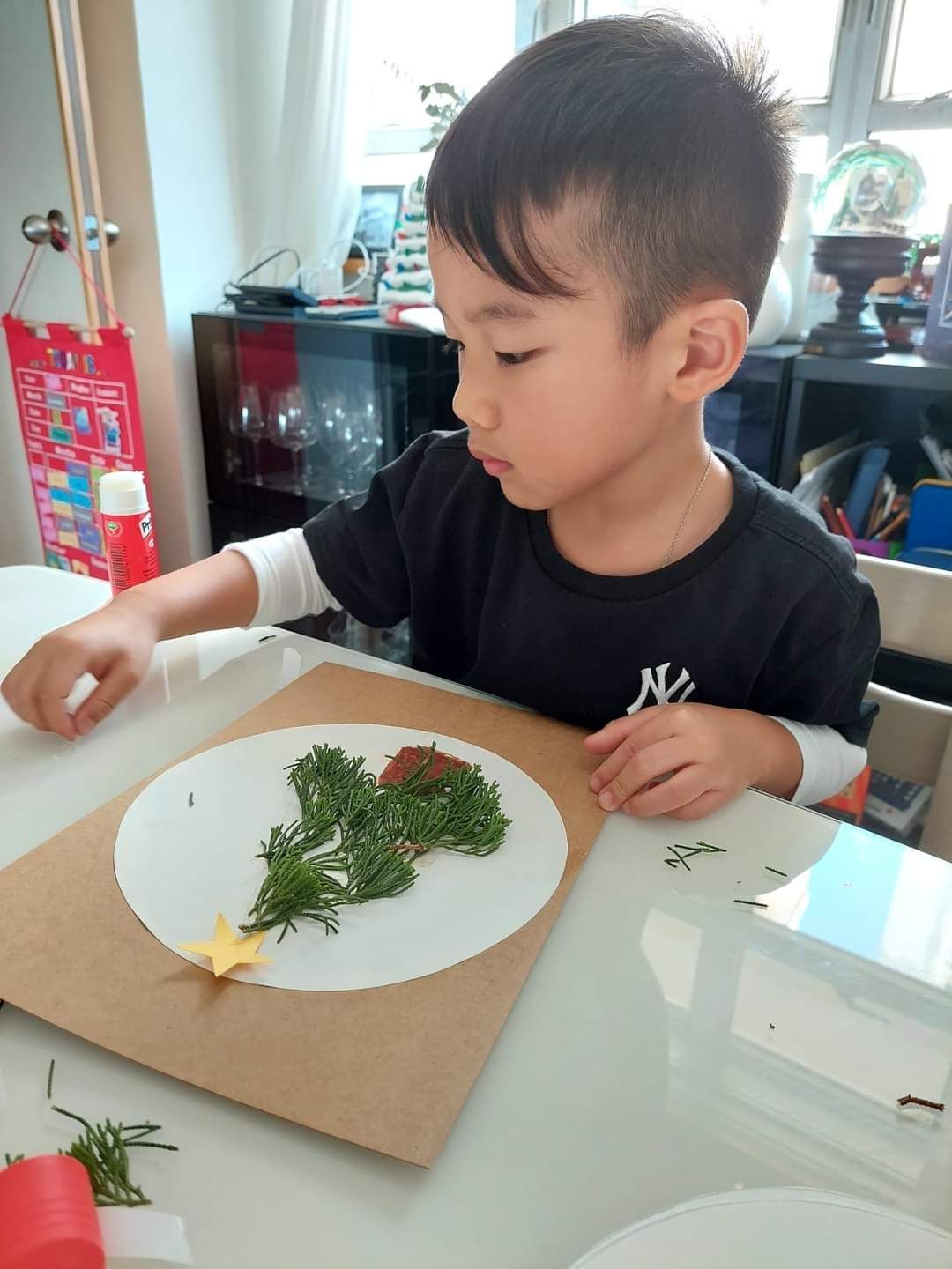 A young boy is sitting at a table making a picture of a christmas tree.