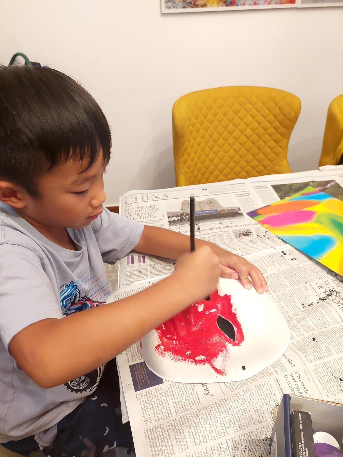 A young boy is painting a paper plate mask with red paint