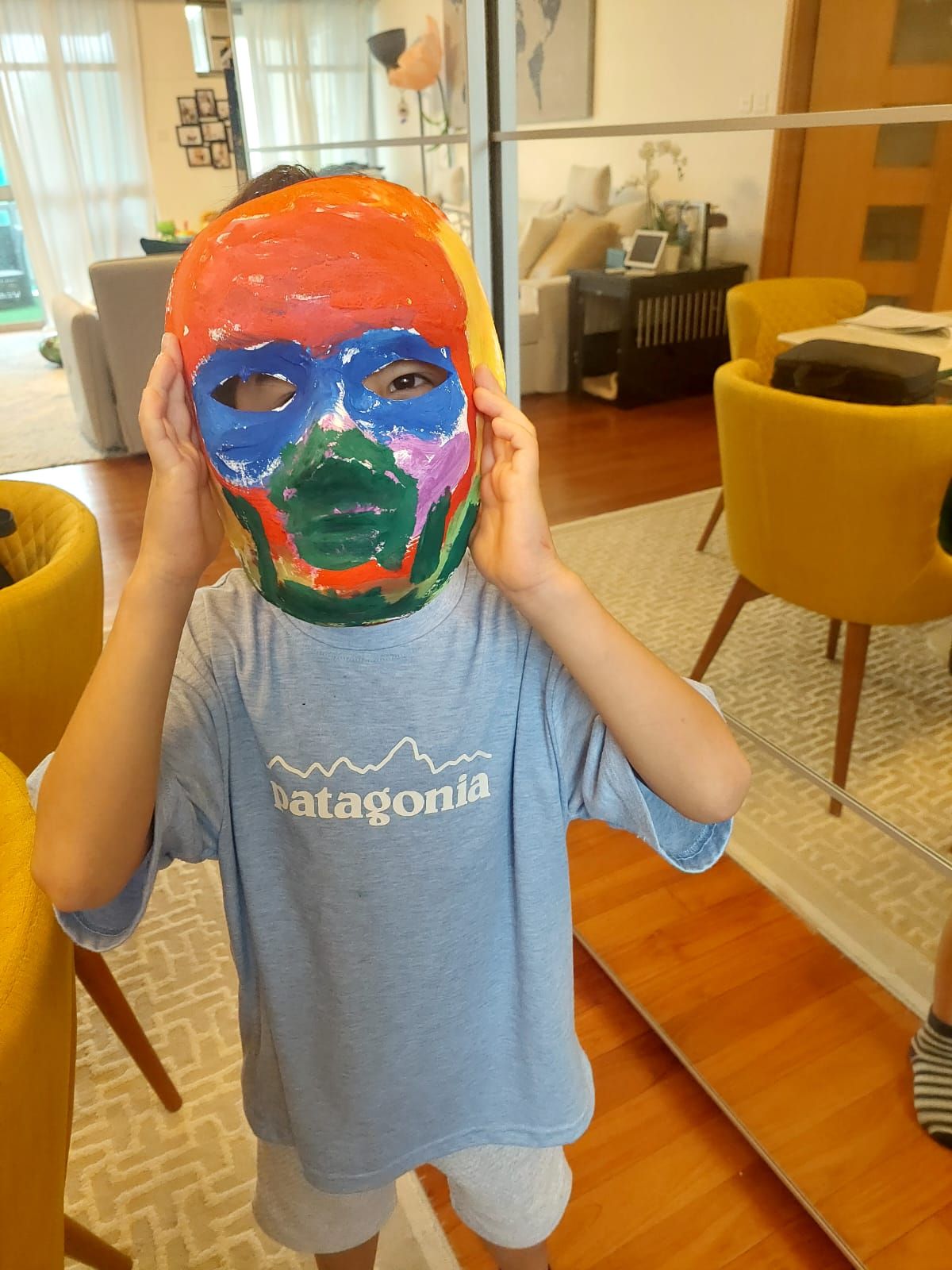 A young boy wearing a patagonia shirt is holding a colorful mask on his face.