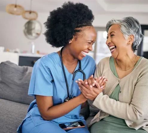 A nurse and an older woman are sitting on a couch laughing.
