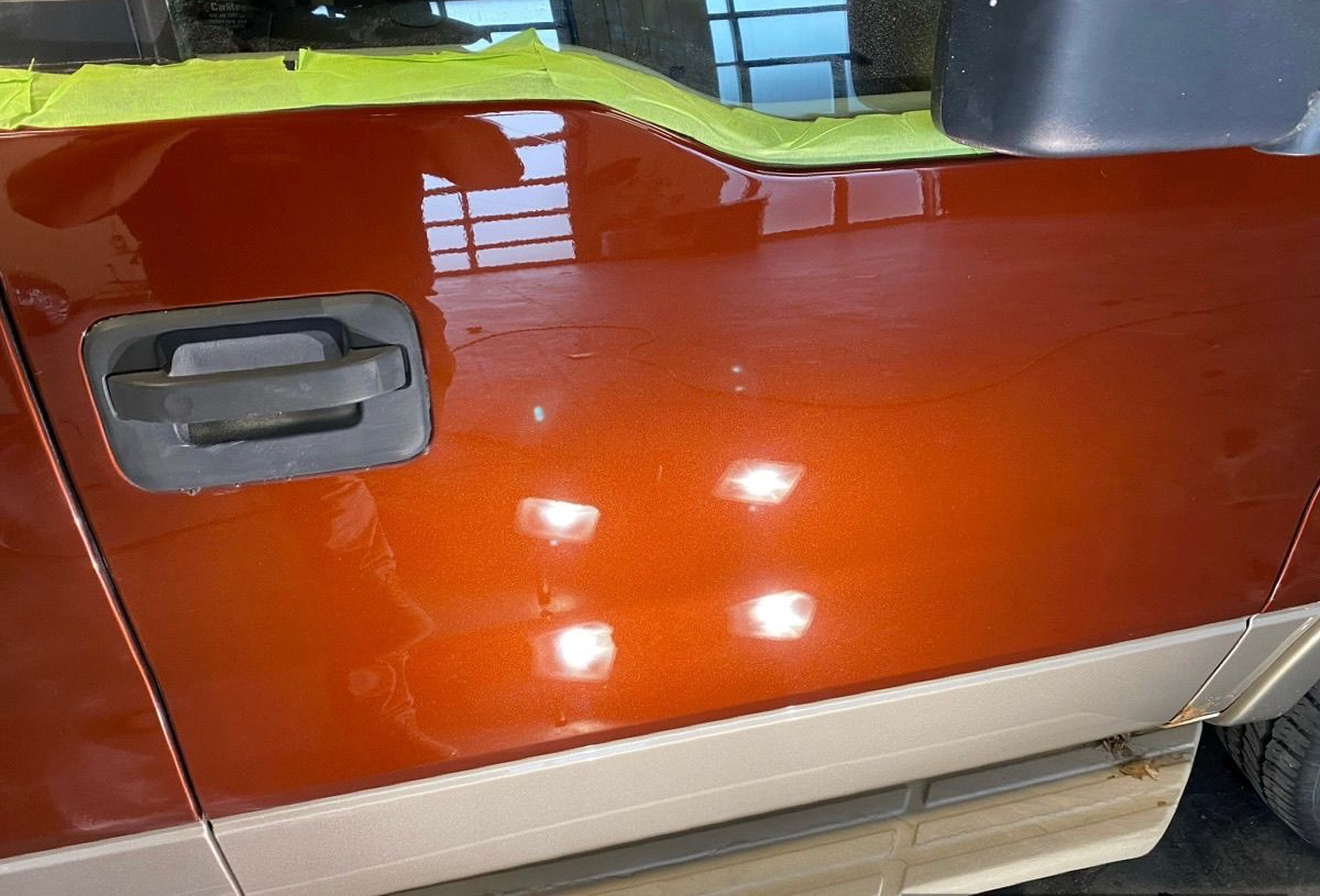 A metallic burnt-orange truck door with a dark handle, beige lower trim, and protective green tape along the window sill.