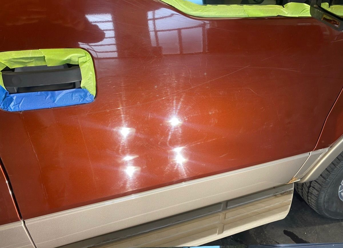 A close-up of a brown car door panel showing surface swirl marks and scratches, with green painter's tape on the handle.
