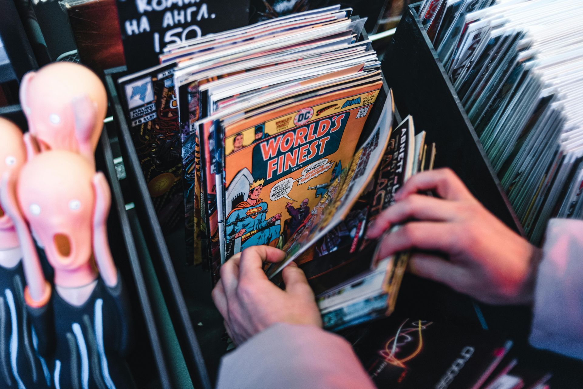 Hands browsing through a rack of comic books in a shop, beside a small figurine resembling a screaming face.