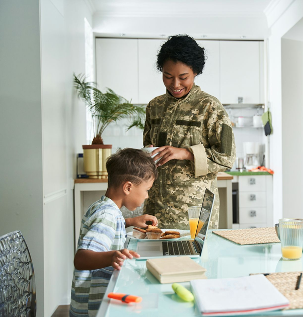 Woman in camo uniform smiles at child at table. Kitchen setting.