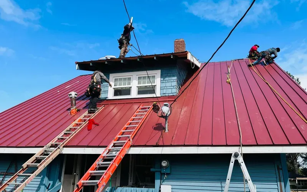 Roofing crew installing red standing-seam metal roof panels on blue house