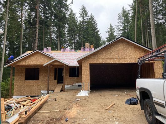 New home construction with roof framing and underlayment installation beginning, surrounded by forest