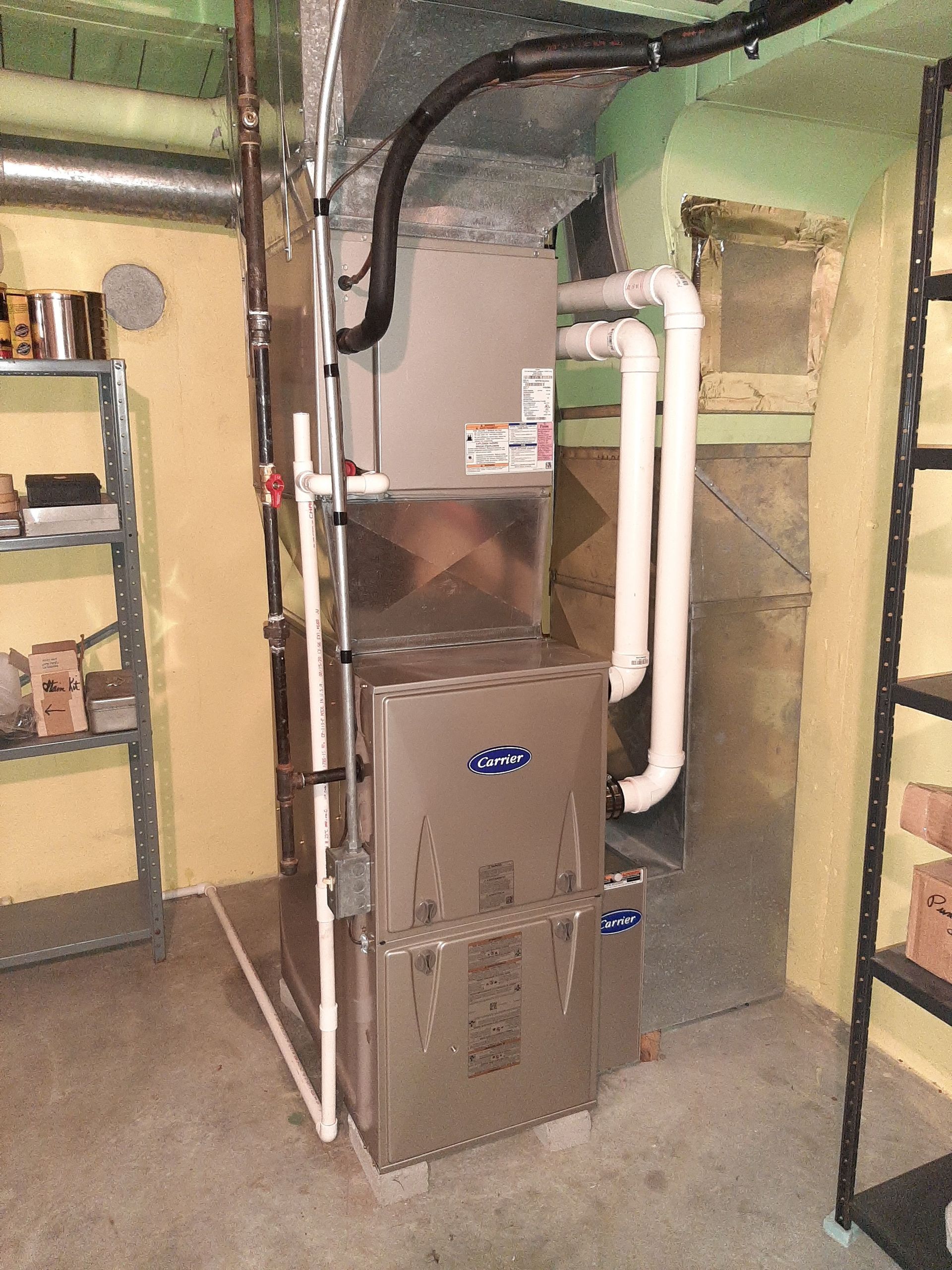 Furnace unit in a basement. Metal structure with white pipes, against a yellow wall, near shelving.