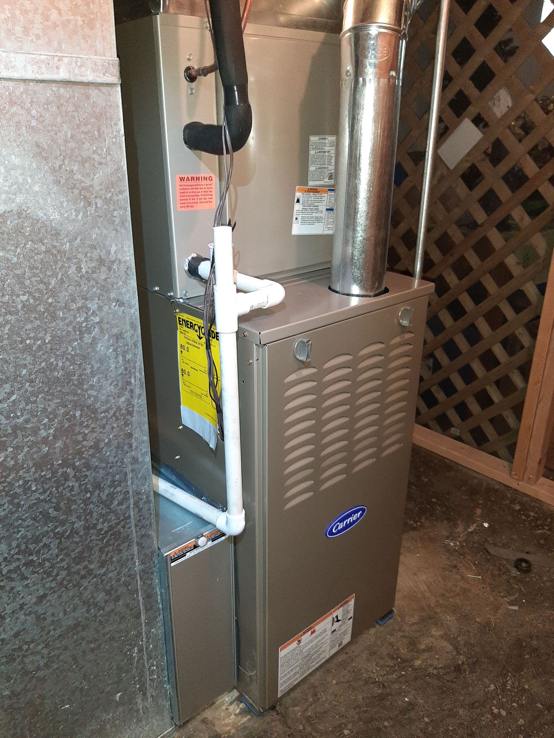 Furnace unit with a silver exhaust pipe and white drainage pipes in a basement.