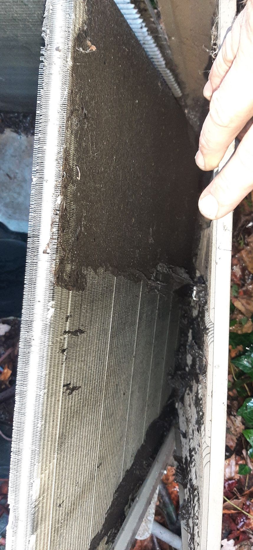 Dirty air conditioning unit. Dark debris covers the cooling fins. A hand holds the edge. Outdoor setting.
