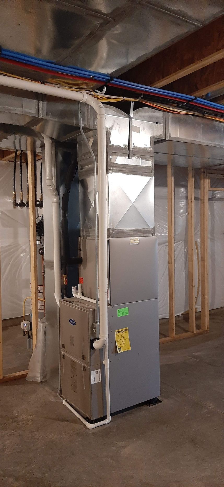 HVAC system installed in a basement, with ductwork and plumbing visible. Gray furnace and wooden framing are present.