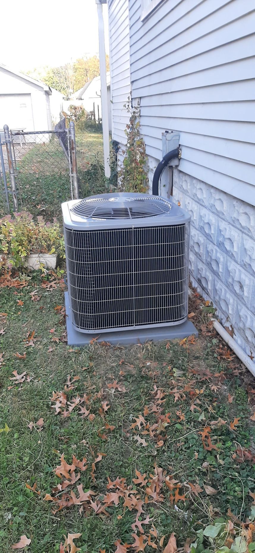 An air conditioning unit sits next to a house on a concrete pad, surrounded by grass and fallen leaves.