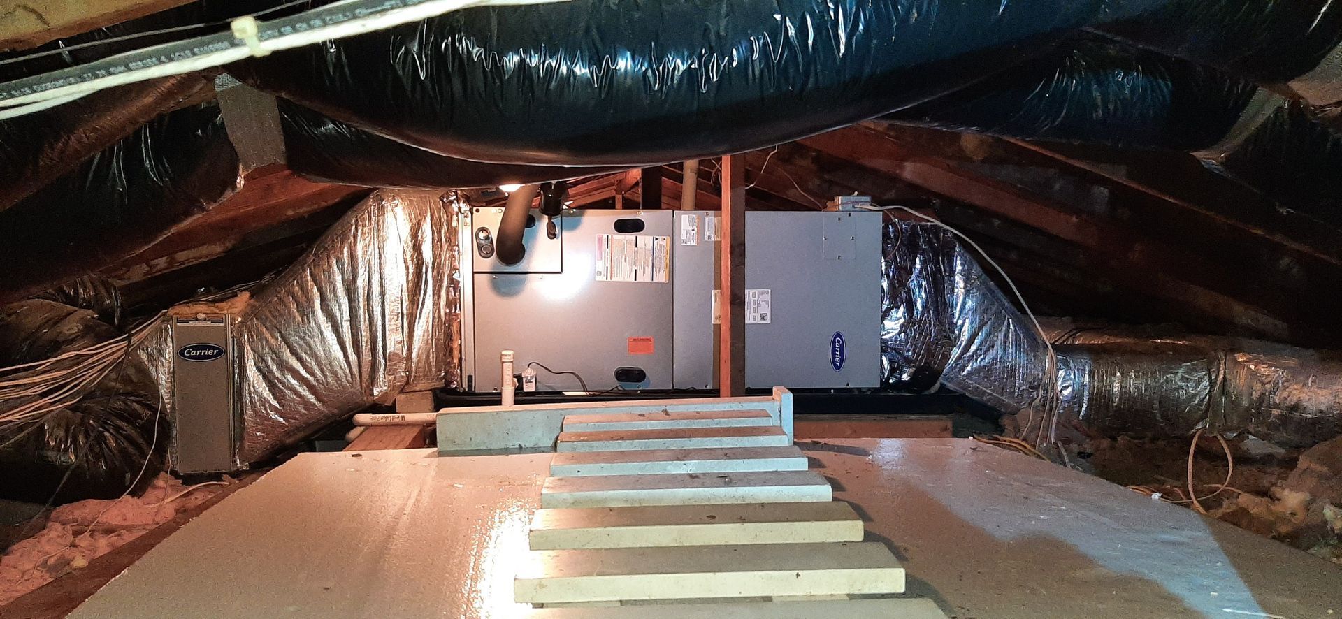 Attic with HVAC unit, ductwork, and wooden steps. Insulation and dark overhead ducts are visible.