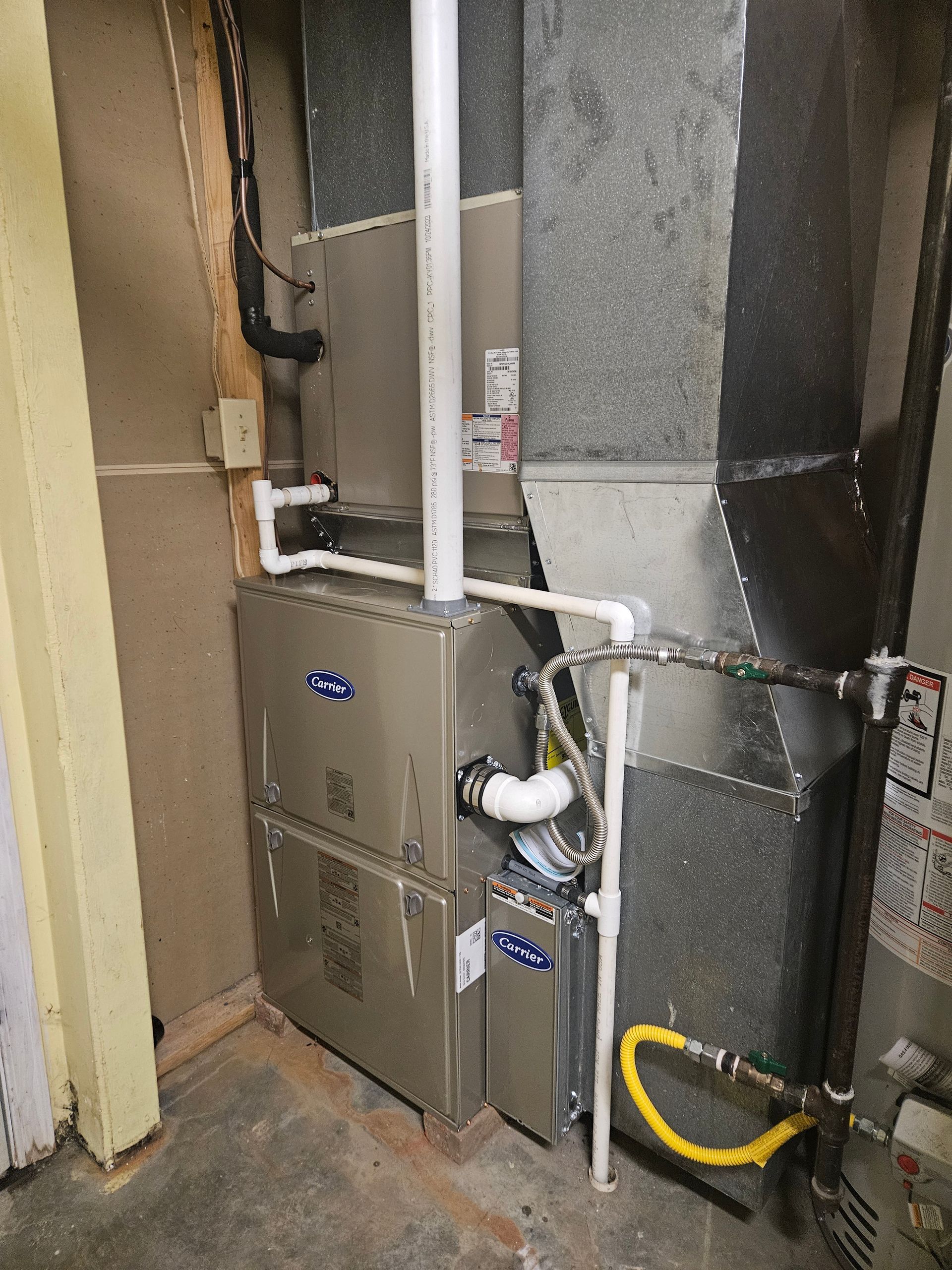 Furnace unit with metallic gray casing and ductwork in a basement setting, connected by white pipes.