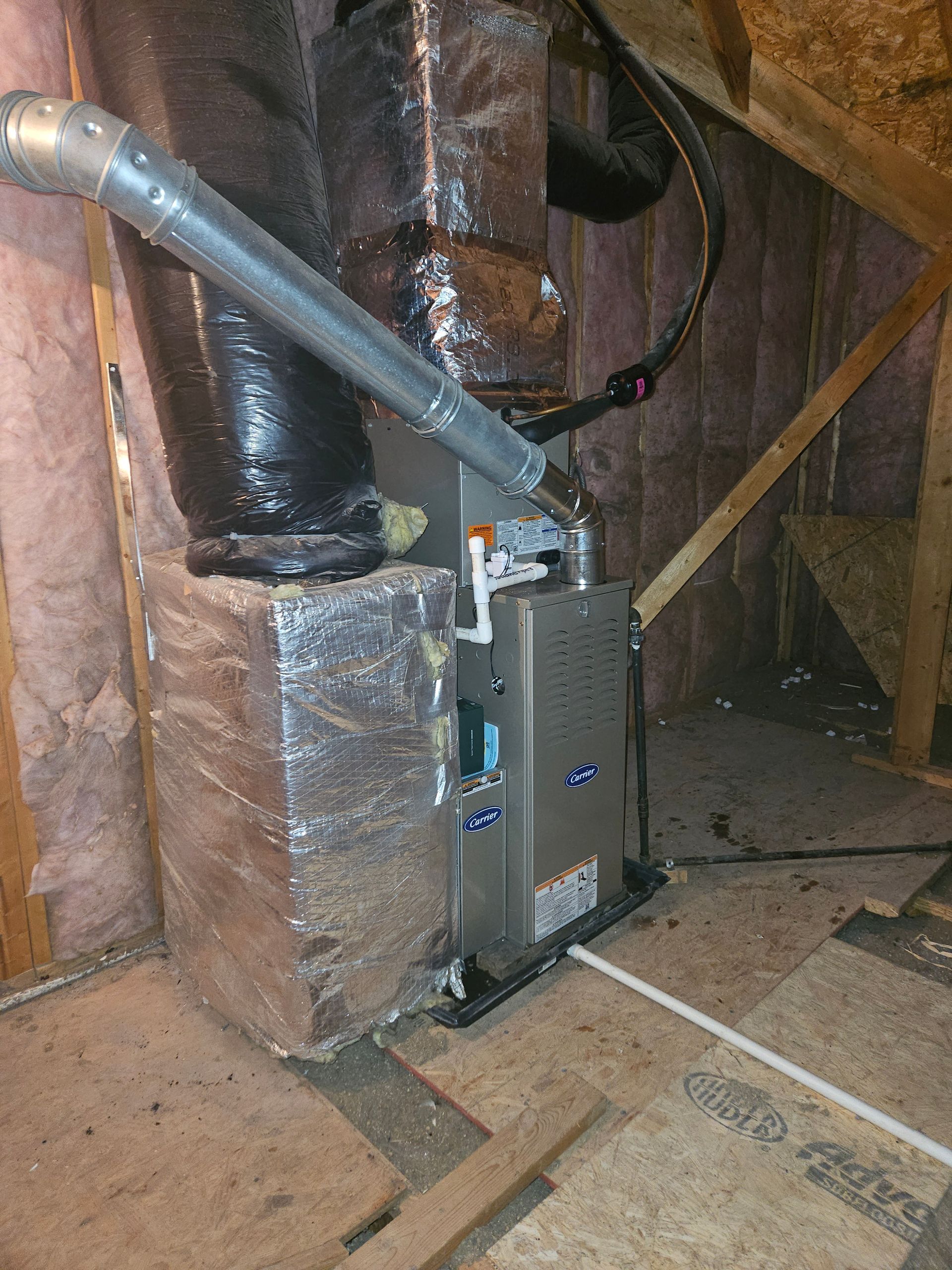 Furnace in an attic with ductwork and insulation. Silver, black, and gray tones in a wood-framed space.
