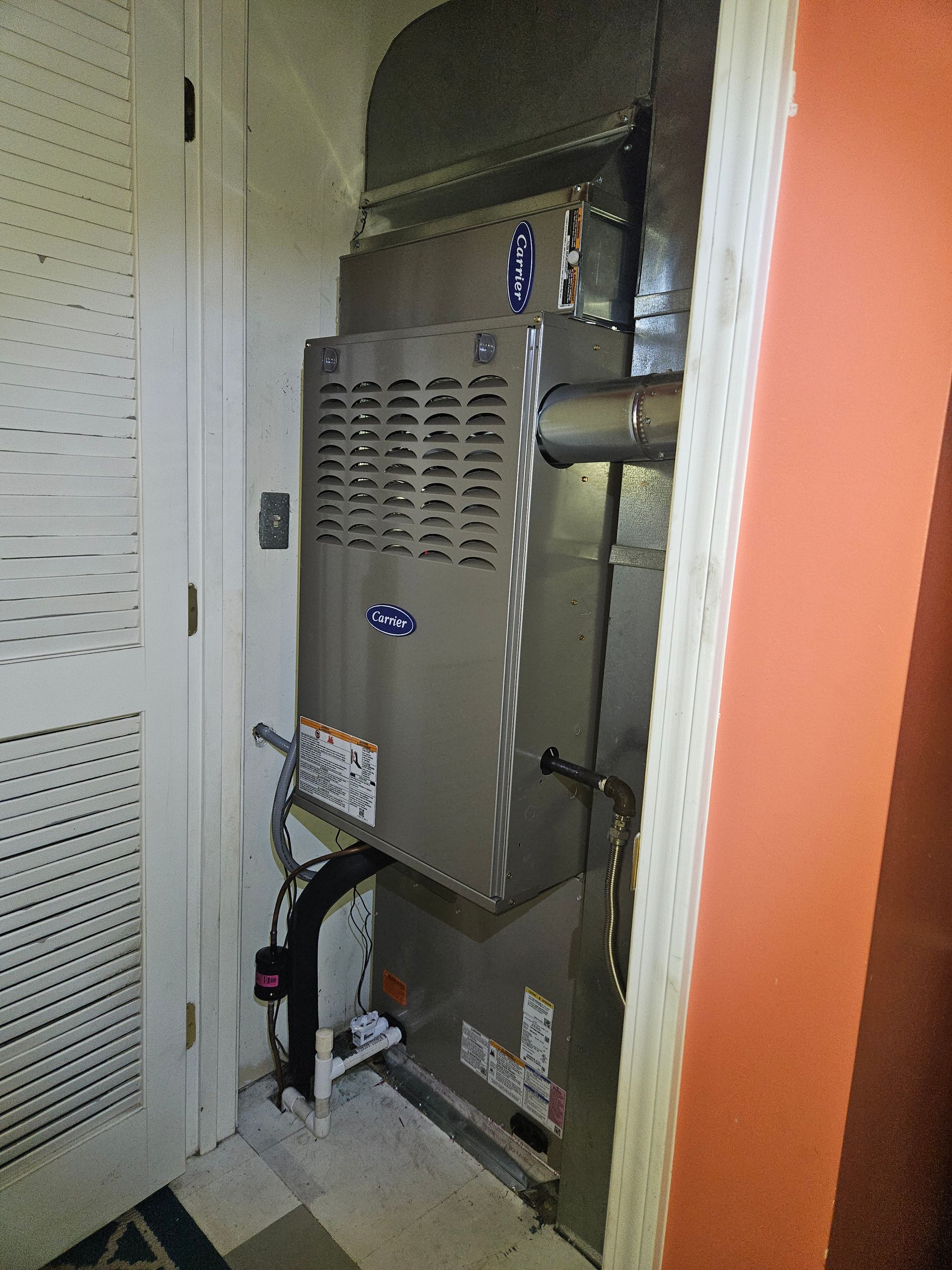 Furnace in a utility closet with a white door and orange wall. Metal ducts and pipes are visible.