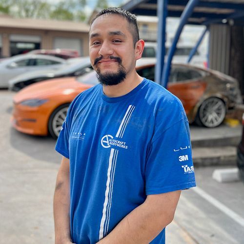 Man in blue shirt smiles in front of orange and damaged cars, likely an auto shop.