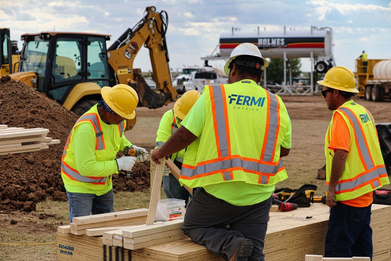 Construction workers in safety vests and helmets building outdoors. Yellow excavator and tanker in background.