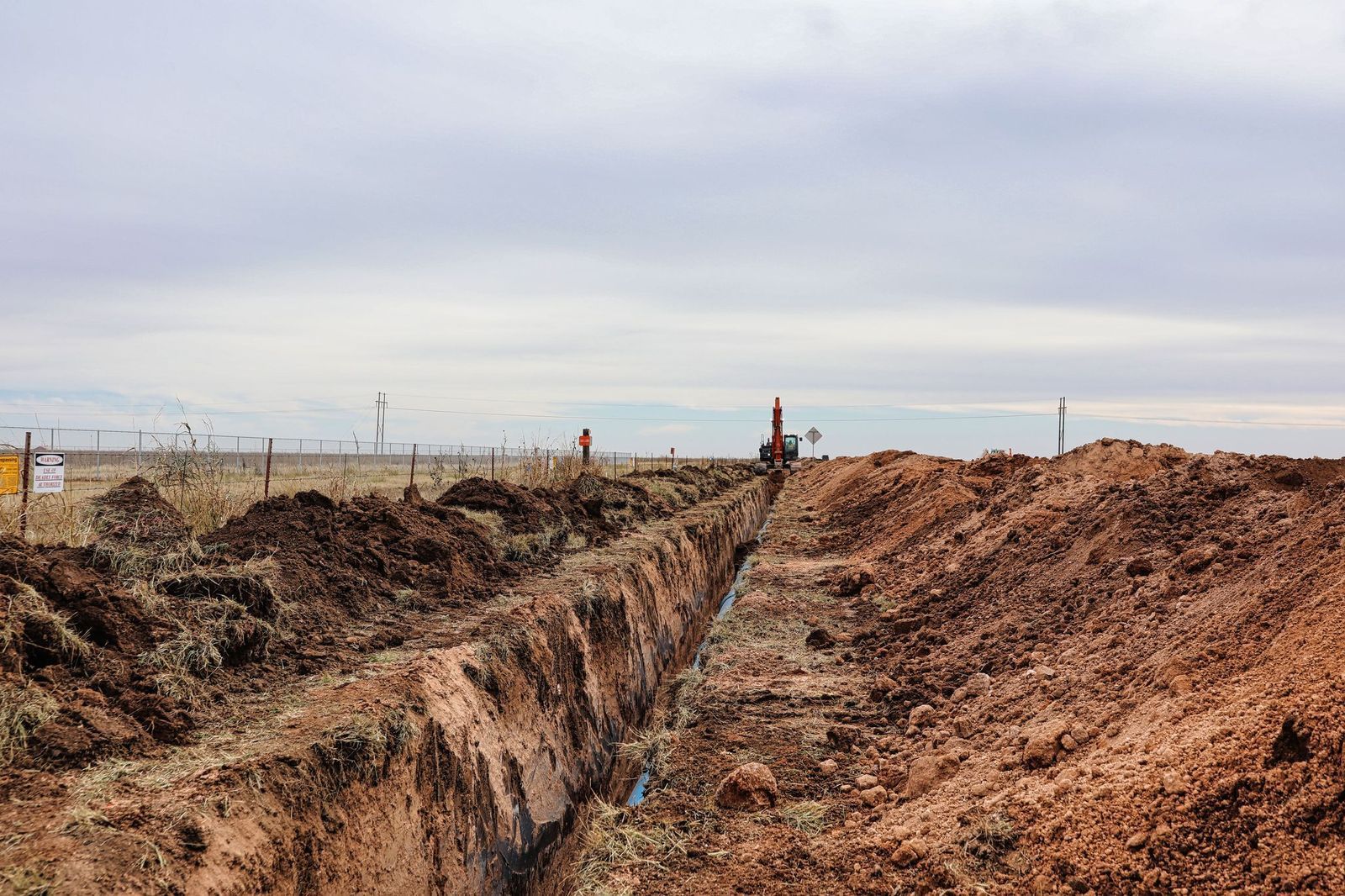 A long, open trench in a field, with piles of reddish-brown dirt on both sides under a cloudy sky.