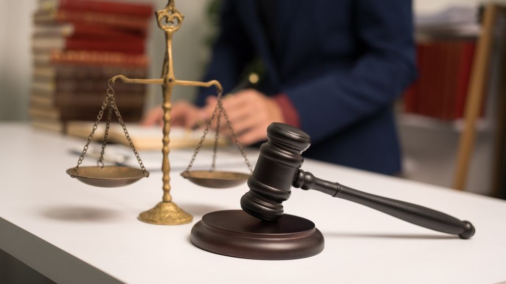 A gavel and scales of justice sit on a white desk with a person in a blue blazer in the background.