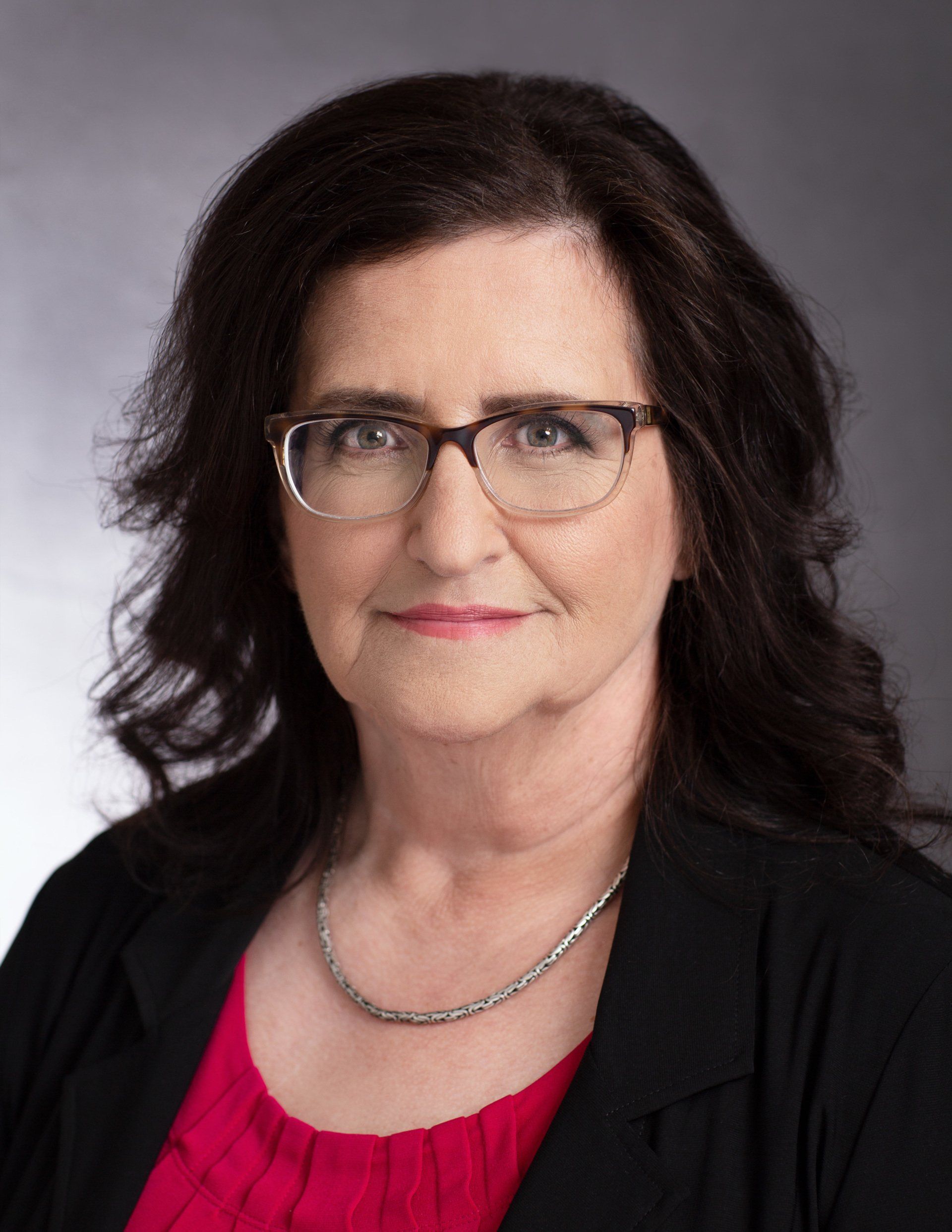 A woman wearing glasses and a red shirt is standing in front of a gray background.