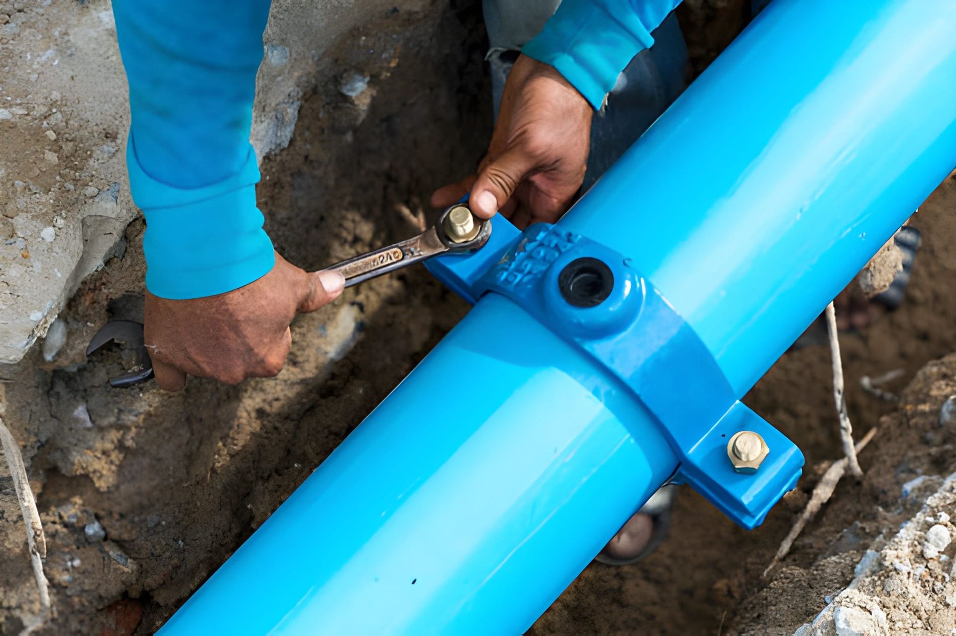 A person using a wrench to tighten bolts on a blue pipe clamp underground.