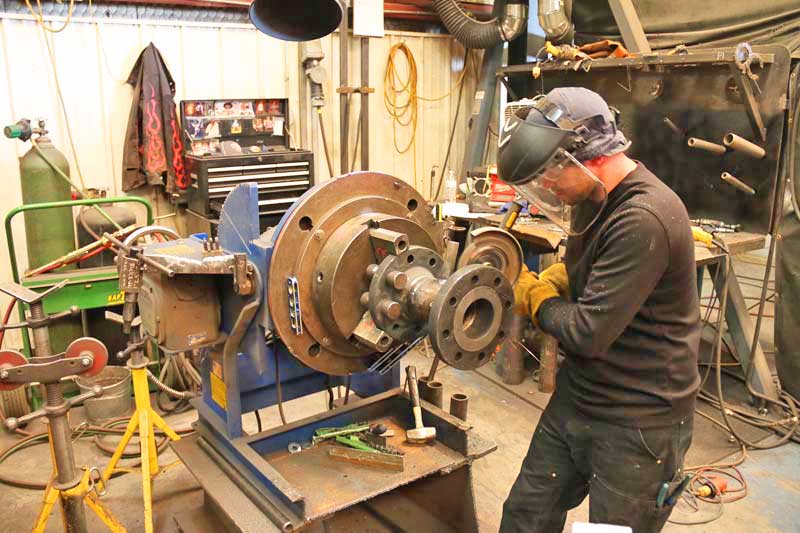 A man wearing a welding mask is working on a machine in a workshop.
