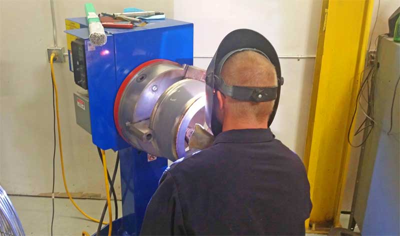 A man wearing a welding mask is working on a machine.