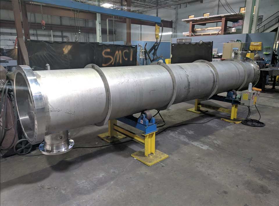 A large stainless steel pipe is sitting on a table in a factory.