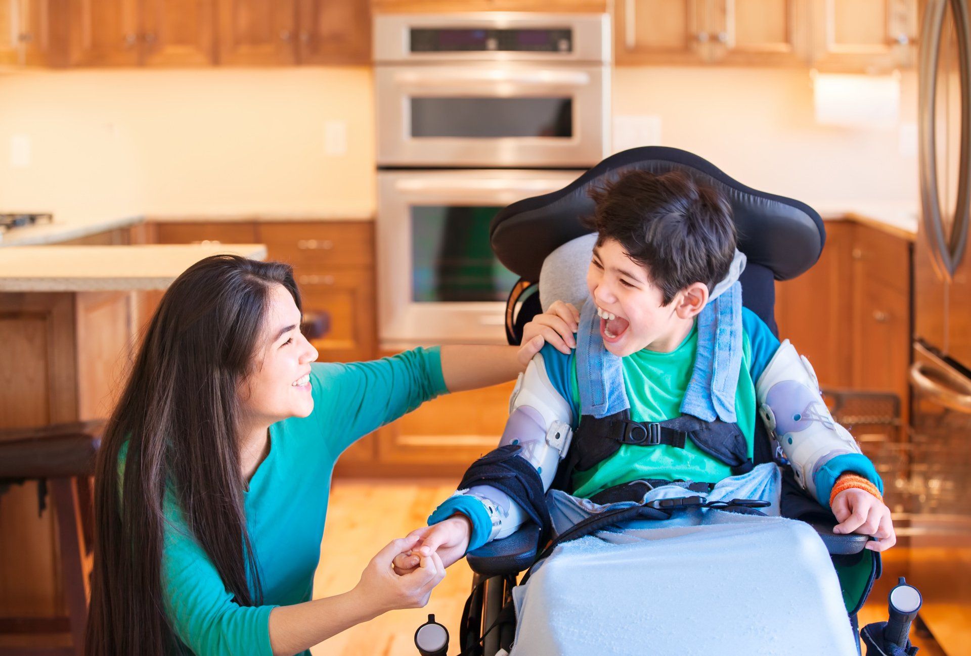 Woman holds hands with laughing boy in a wheelchair in a kitchen.