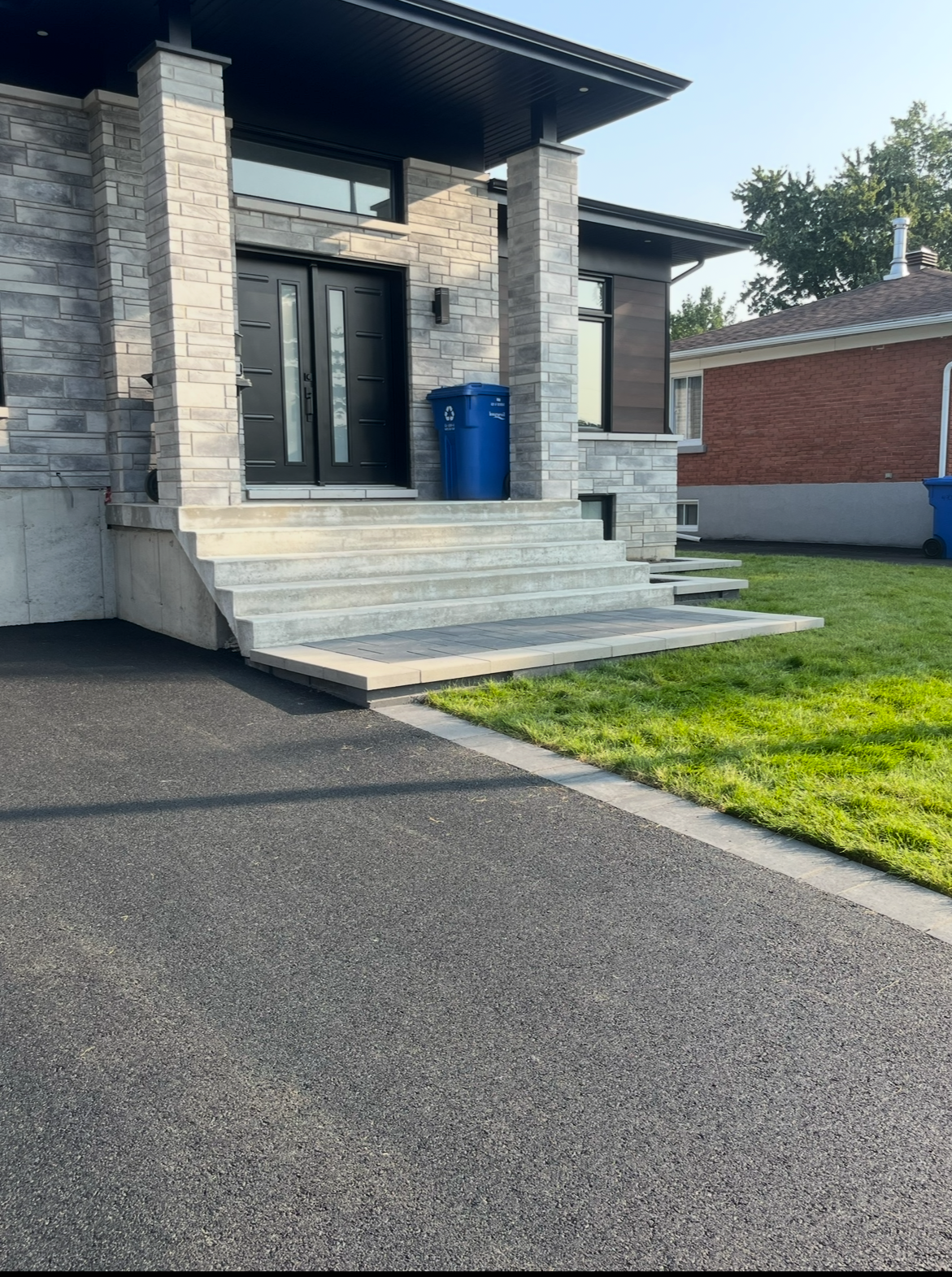 Une maison avec une allée noire et une poubelle bleue devant.