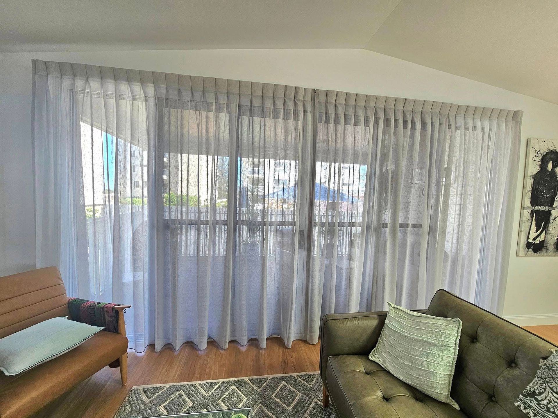 Living room with sheer grey curtains over a large window, two couches, and a rug.  A painting hangs on the wall. — Modern Window Furnishings In Childers, QLD