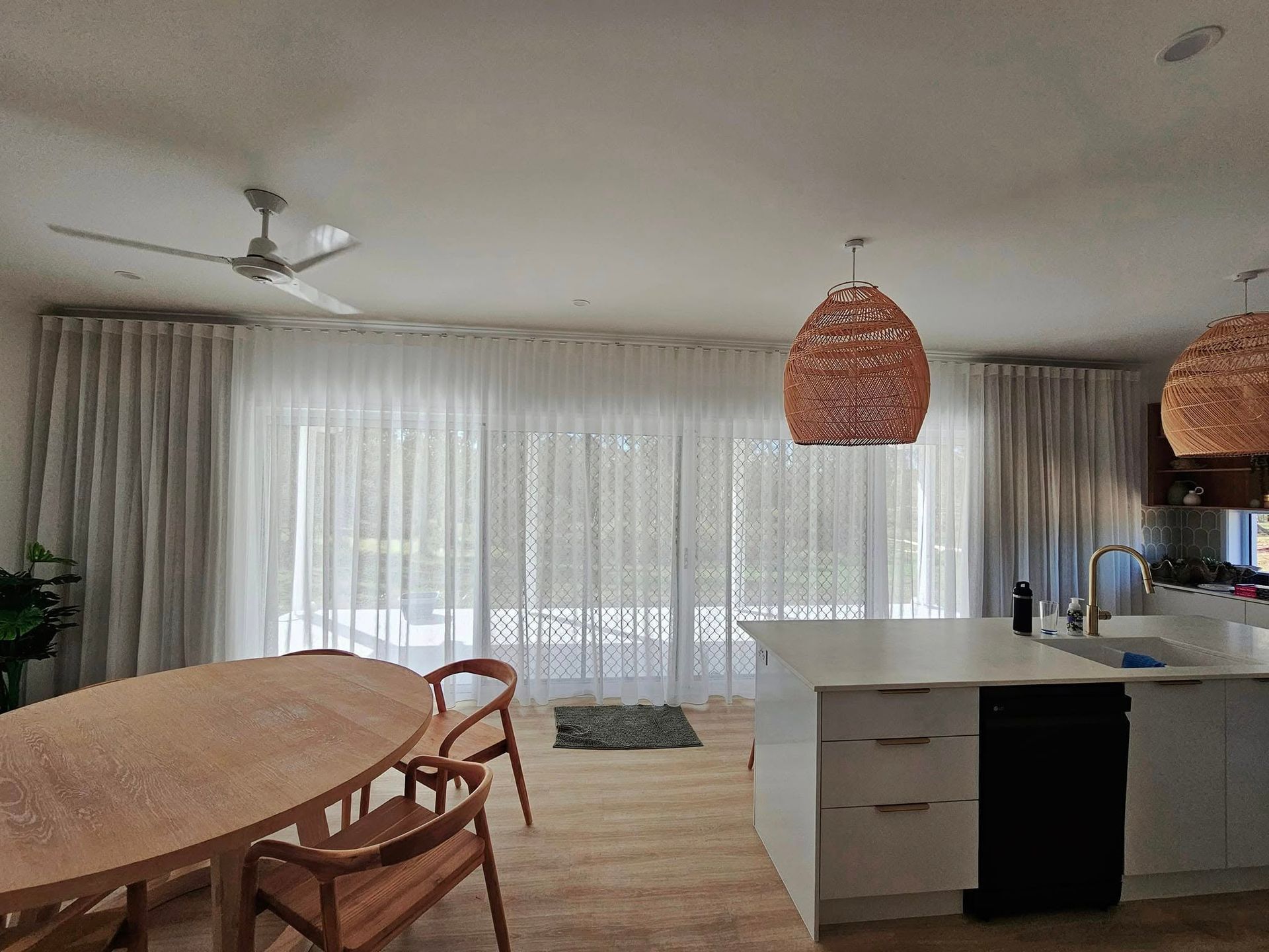 A bright dining room with large windows covered by sheer curtains. A wooden table and chairs sit in the foreground, and a kitchen island with pendant lights is visible. — Modern Window Furnishings In Bargara, QLD