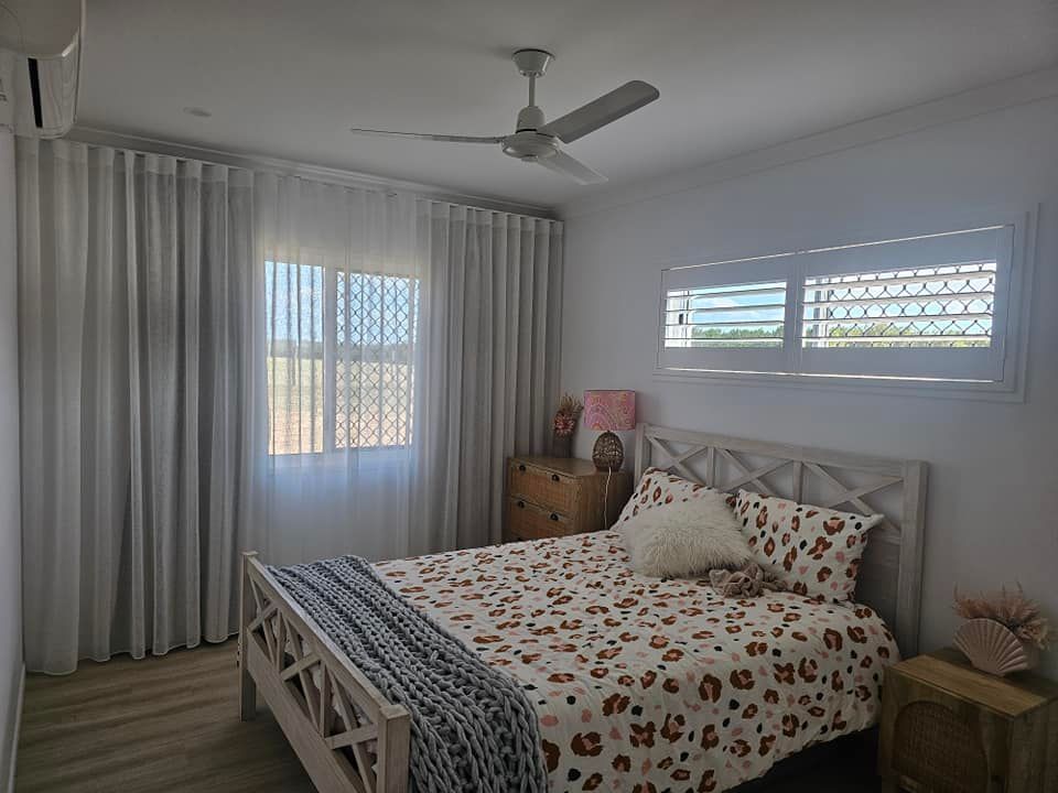 Bedroom with a bed, two windows with coverings, and two wooden bedside tables. One window has white shutters, the other has a screen. — Modern Window Furnishings In Gin Gin, QLD