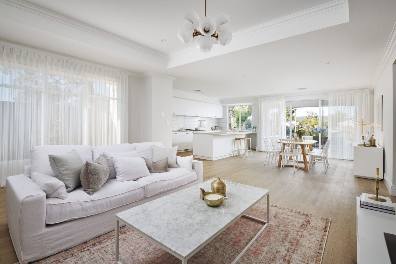 A living room with two windows covered by beige curtains. A chaise lounge and small wooden cabinet are in front of the windows. — Modern Window Furnishings In Bundaberg East, QLD