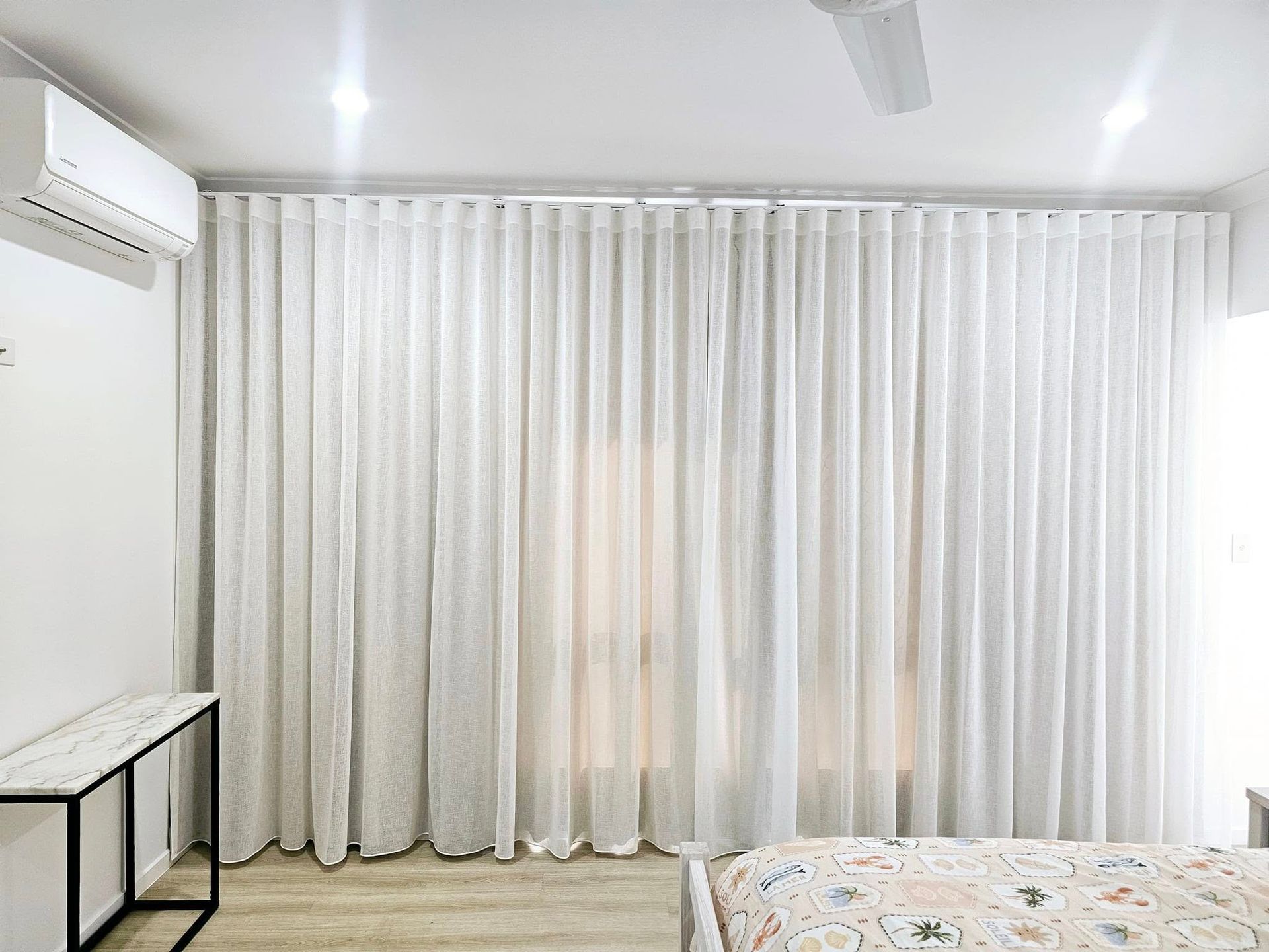 Sheer white curtains covering a corner window in a bedroom. Sunlight streams through, partially illuminating a bed with dark bedding and pillows. — Modern Window Furnishings In South Kolan, QLD