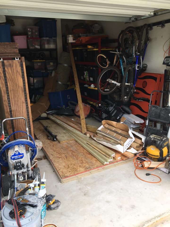 Cluttered garage interior. Paint sprayer, lumber, bikes, and various items stored. Concrete floor, overhead lighting.
