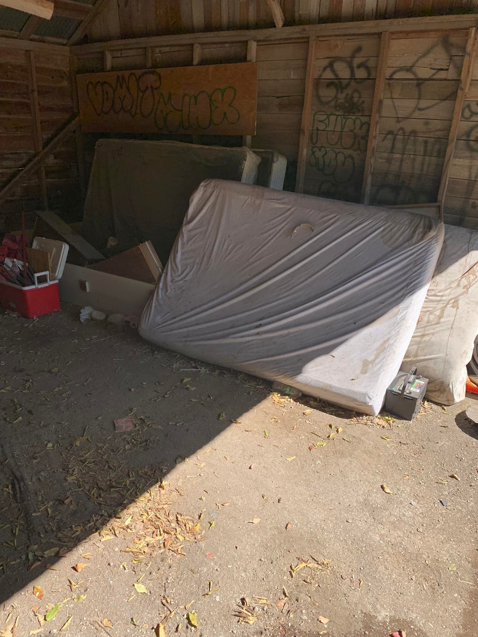 A mattress wrapped in plastic rests inside a shed with other discarded items.