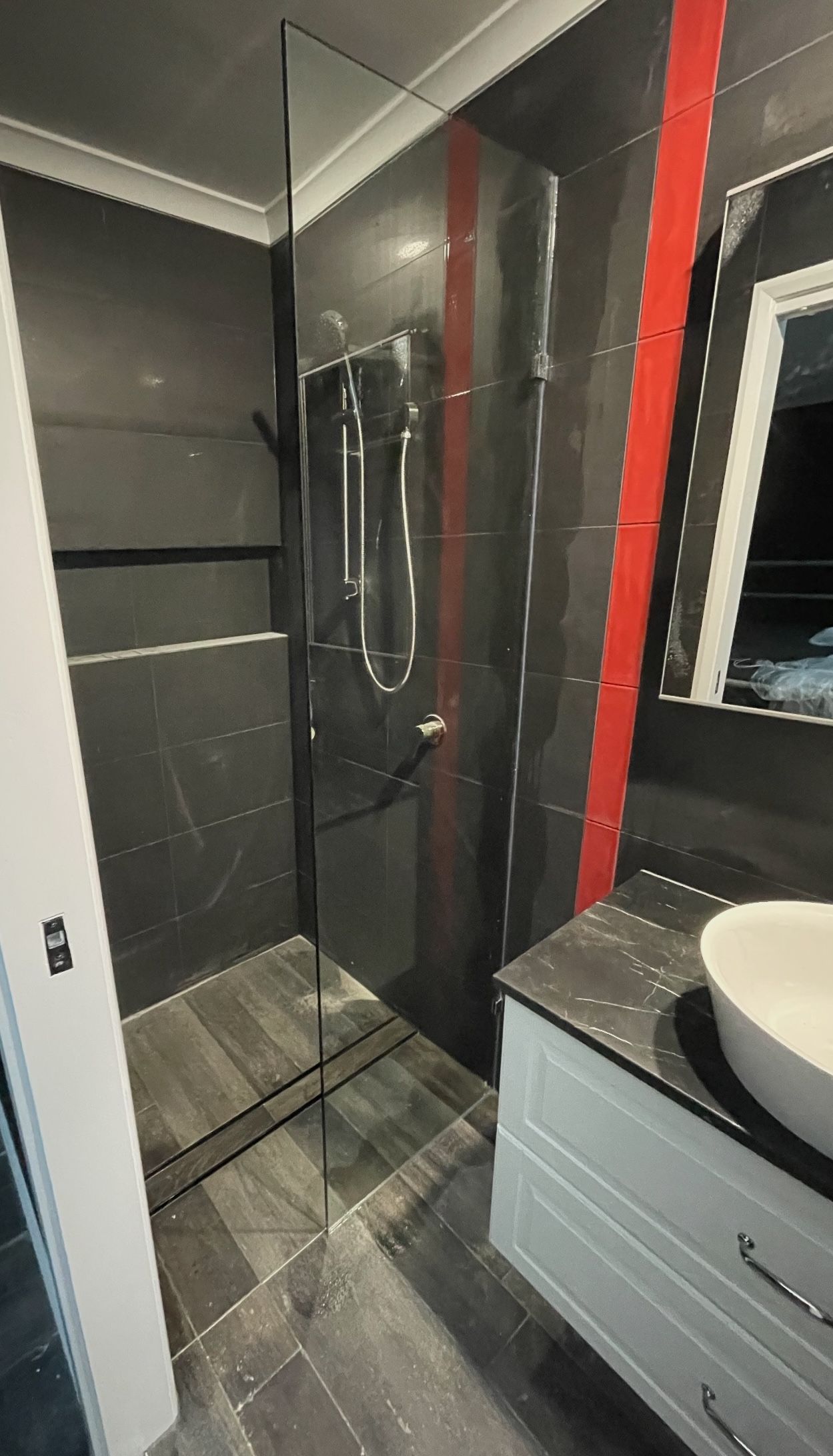 A bathroom with a black tiled walk in shower , sink and mirror.