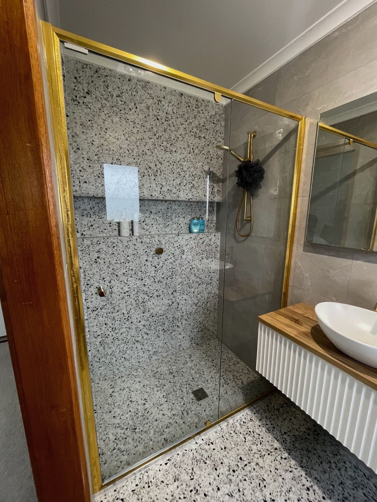 A bathroom with a walk in shower with gold accents , sink and mirror.