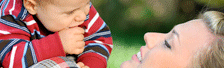 A baby in a red striped shirt looks down while a woman gazes up at him in a grassy outdoor setting.