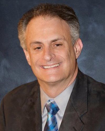 Man in suit smiles, looking at the viewer. Dark blazer, blue patterned tie, gray shirt.