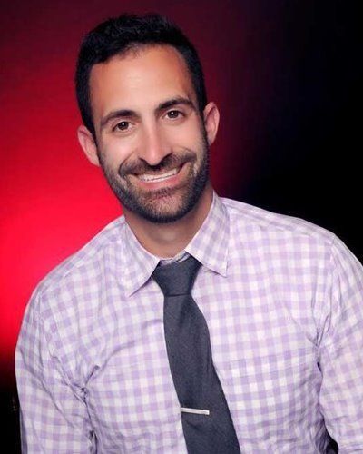 Man smiling, wearing a checked shirt, tie, and tie clip, against a red and black background.