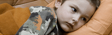A child in bed looking away with a concerned expression. A camouflage blanket and orange pillow.