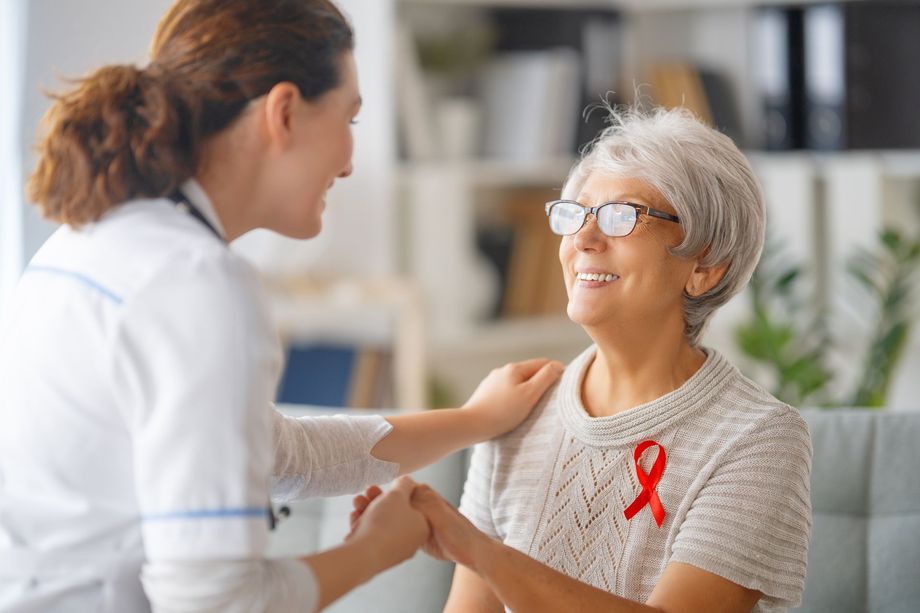 A nurse is holding the hand of an elderly woman with a red ribbon on her shirt.