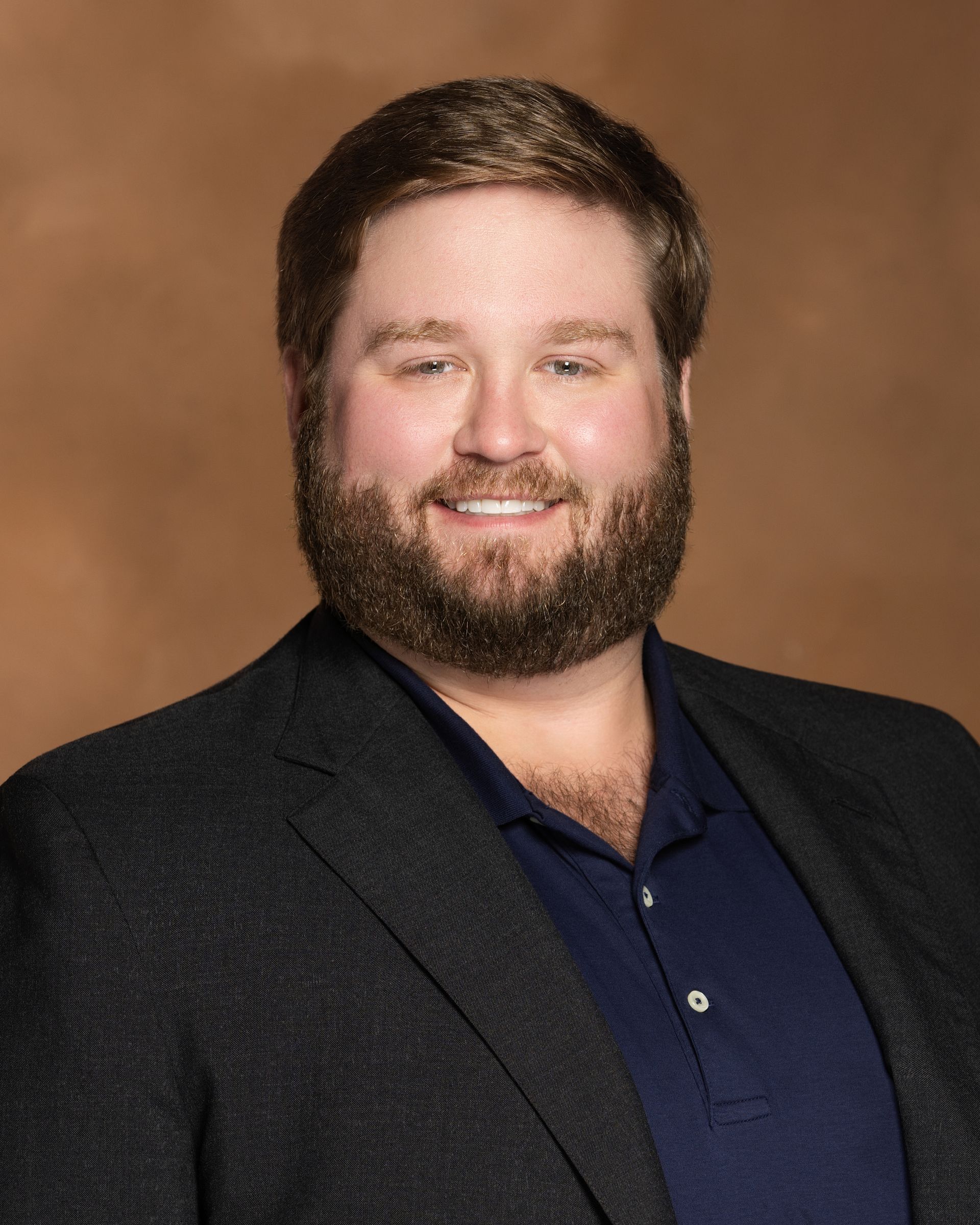 Man in blazer, smiling, dark beard, blue polo shirt.