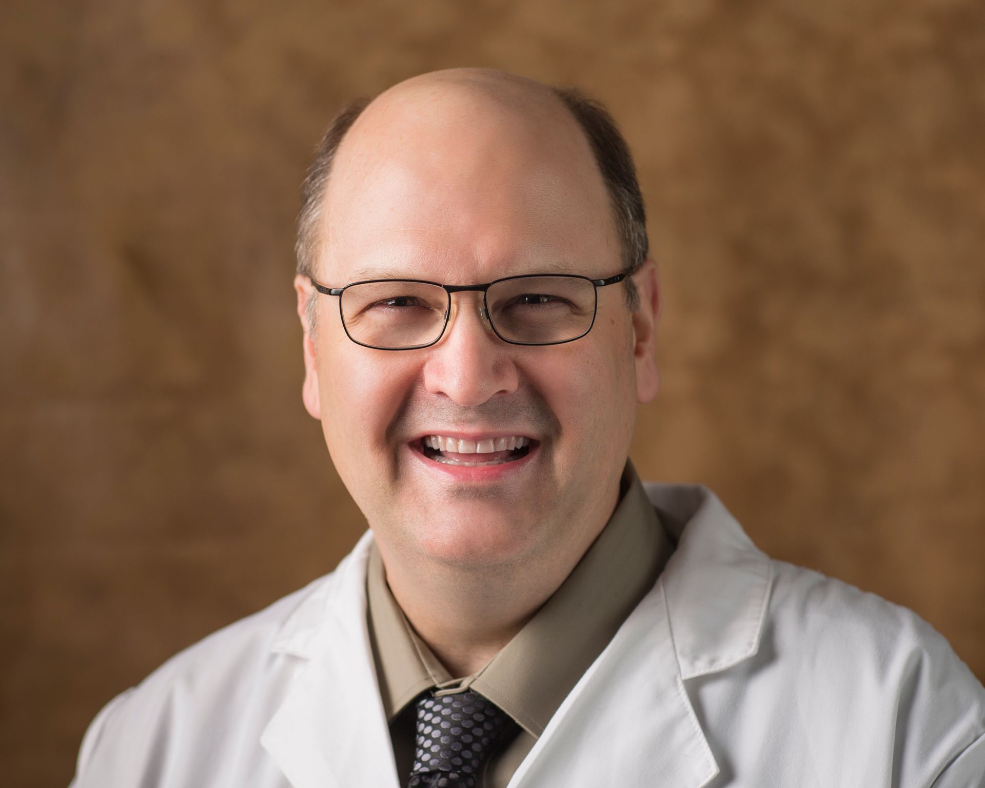 A bald man wearing glasses and a white coat smiles for the camera