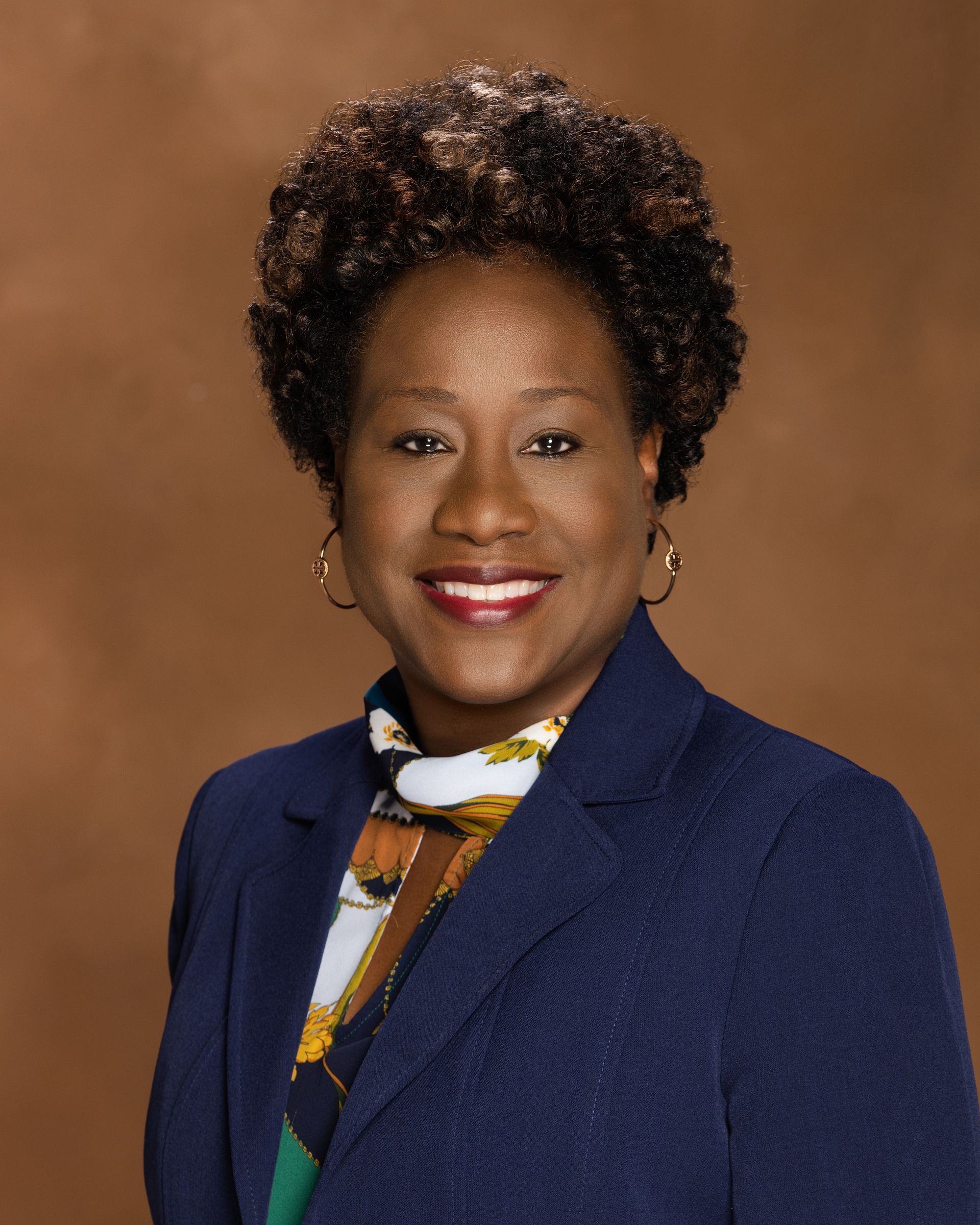 Woman in a blue blazer smiles, wearing gold hoop earrings and a scarf. Brown background.
